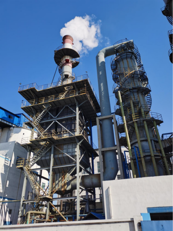 An industrial pelletizing disc machine in a factory, processing dark granular materials, with mechanical components, pipes, and a metal structure in the background.