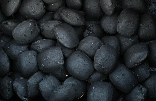 A pile of dark, irregularly shaped briquettes, likely industrial or mineral in nature, with several pairs of shoes visible around, indicating a work or inspection scene.