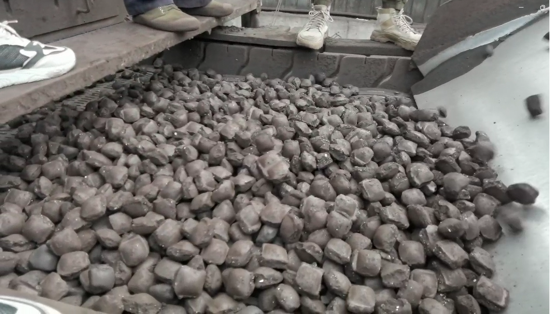A pile of dark, irregularly shaped briquettes, likely industrial or mineral in nature, with several pairs of shoes visible around, indicating a work or inspection scene.