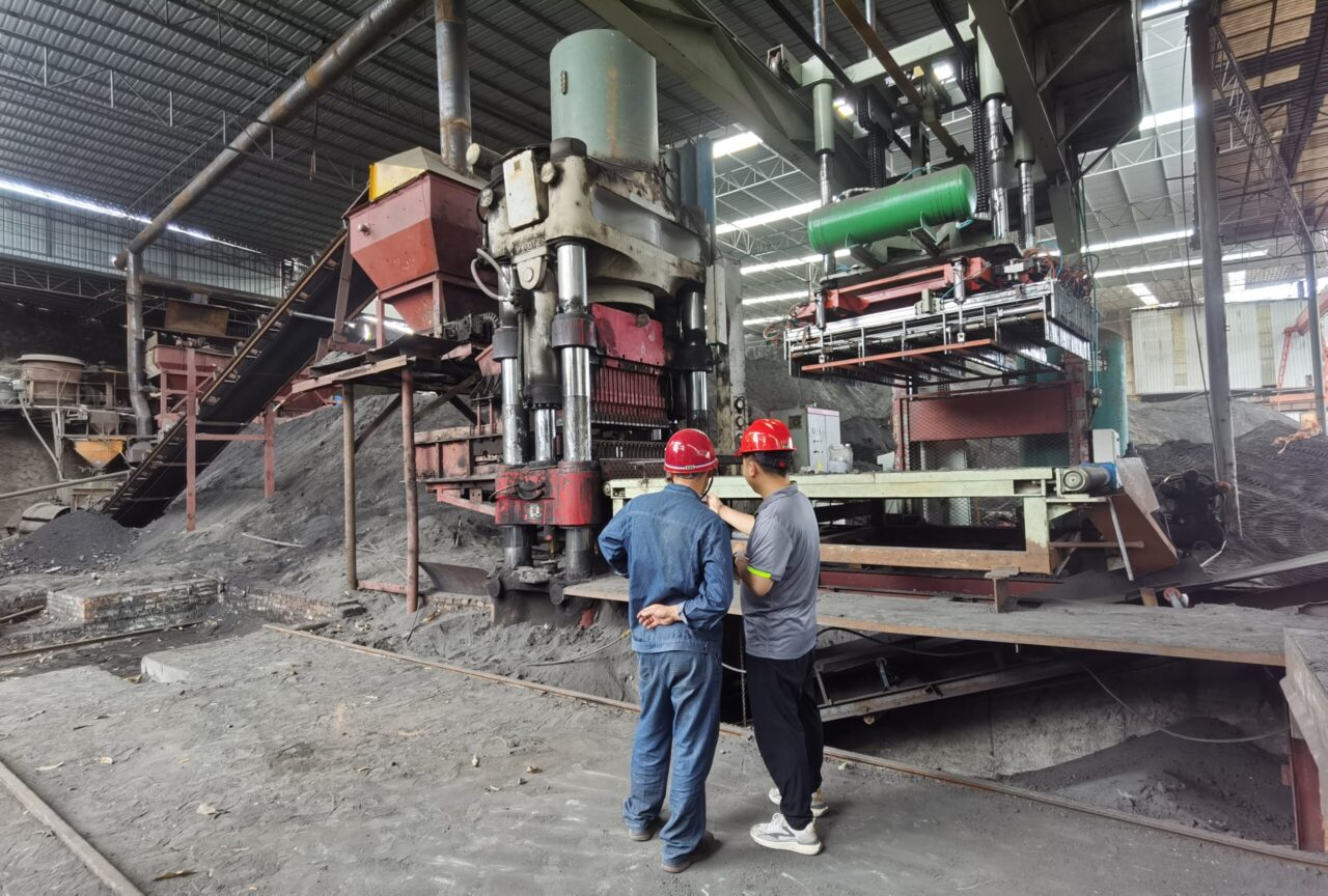 An industrial pelletizing disc machine in a factory, processing dark granular materials, with mechanical components, pipes, and a metal structure in the background.